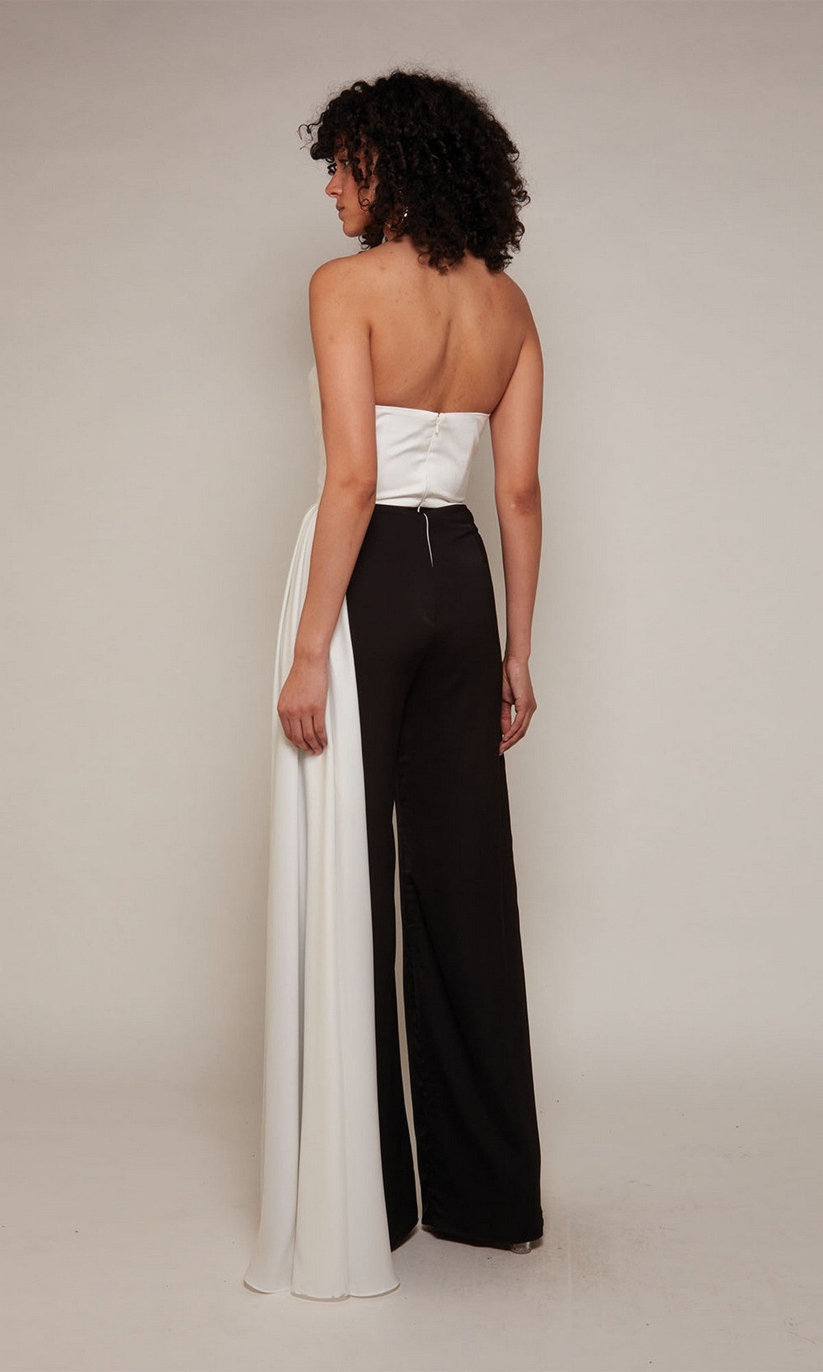 A person stands facing away, wearing the Long Formal Jumpsuit: Alyce 70050—an elegant strapless style with a white top, black pants, and a draped white panel on one leg—set against a plain light background.